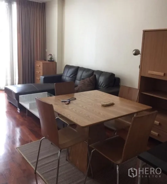 Condominium for rent or sale in Khlong Toei, Bangkok - Cozy living room with black sectional sofa, wooden dining set, and large curtained window.
