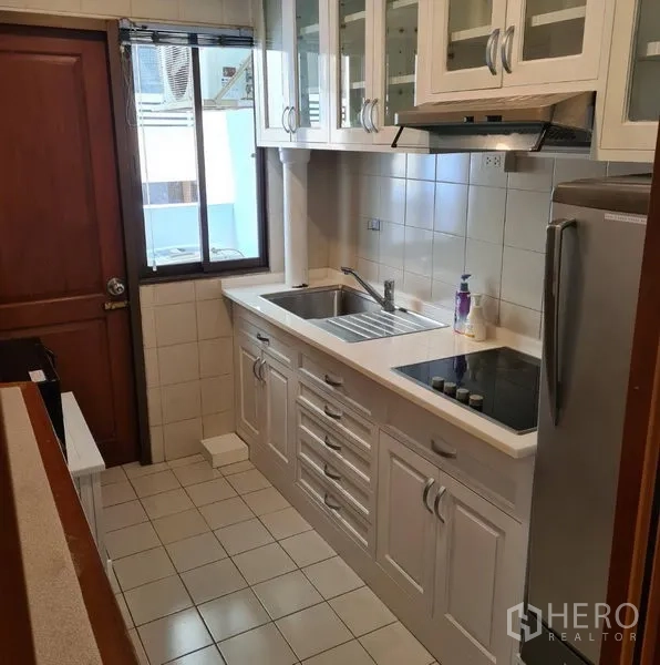 Condominium for rent or sale in Watthana, Bangkok - Compact galley kitchen with white cabinetry, glass-front cupboards, electric cooktop, sink and window ventilation.