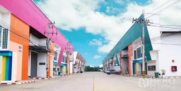 Warehouse for rent in Mueang Samut Sakhon, Samut Sakhon - Row of modern, colorful warehouse units with wide driveway, roller shutter doors, and overhead power lines in an industrial complex.
