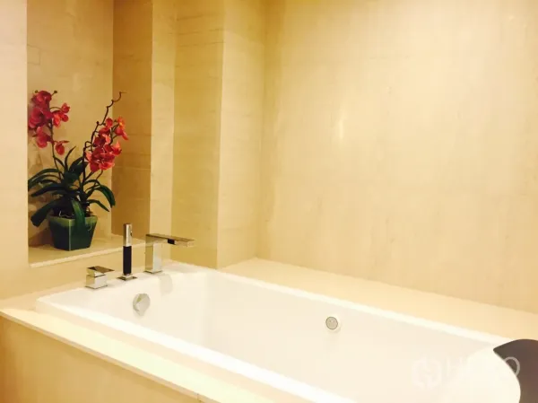Condominium for rent or sale in Sathon, Bangkok - Deep soaking bathtub set in beige stone with chrome fixtures and a decorative orchid.