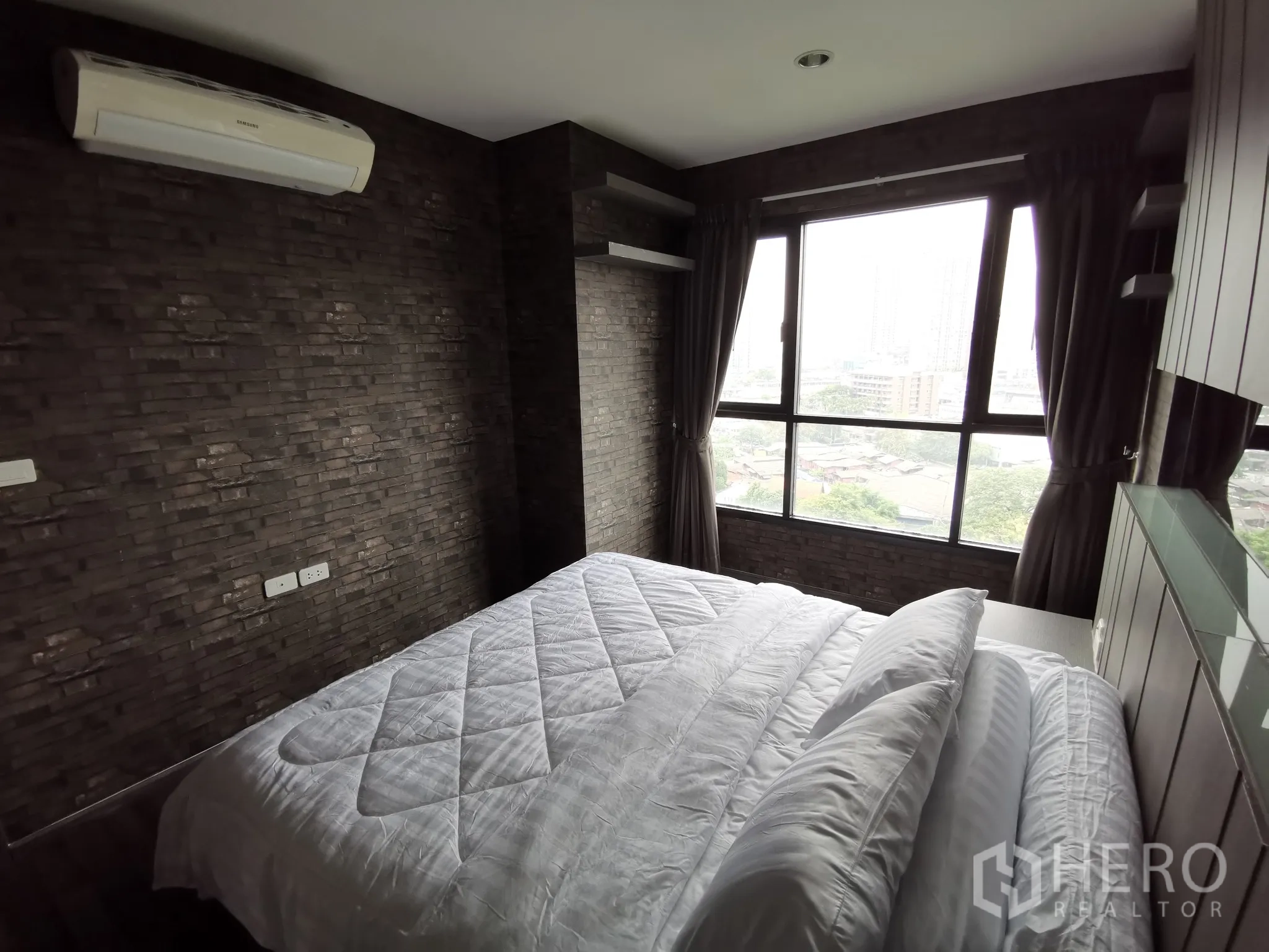 Condominium for rent or sale in Watthana, Bangkok - Master bedroom showing two windows, dark brick accent walls and city view.
