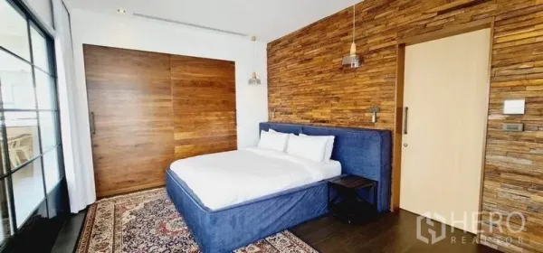 Condominium for rent in Watthana, Bangkok - Bedroom with textured wood accent wall, sliding timber door, upholstered bed and rug.