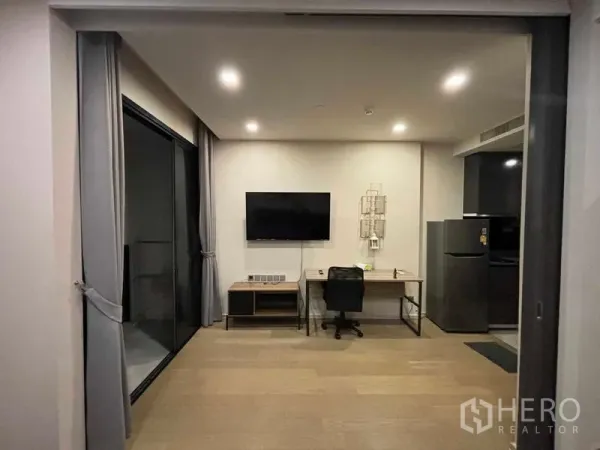 Condominium for rent in Watthana, Bangkok - Open-plan living area with wall-mounted TV, work desk, balcony sliding doors and recessed lighting.