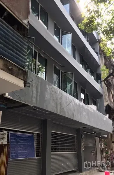 Shophouse for rent in Khlong Toei, Bangkok - Newly renovated multi-storey shophouse facade with grey exterior, glass windows and ground-floor roller shutters on a tree-lined Sukhumvit lane.
