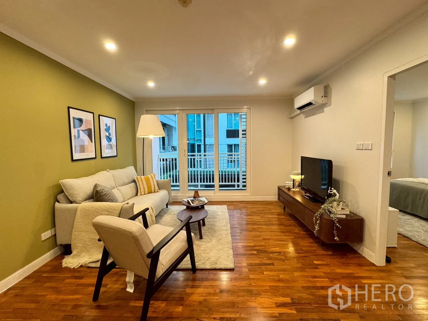 公寓 for sale in Watthana, Bangkok - 客厅配沙发与单椅、电视柜、木地板，并有推拉门通往阳台，旁有卧室门。