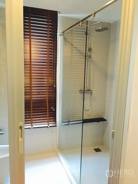 Condominium for rent or sale in Khlong Toei, Bangkok - Contemporary bathroom with glass-enclosed rain shower and bench by a window with wooden blinds.