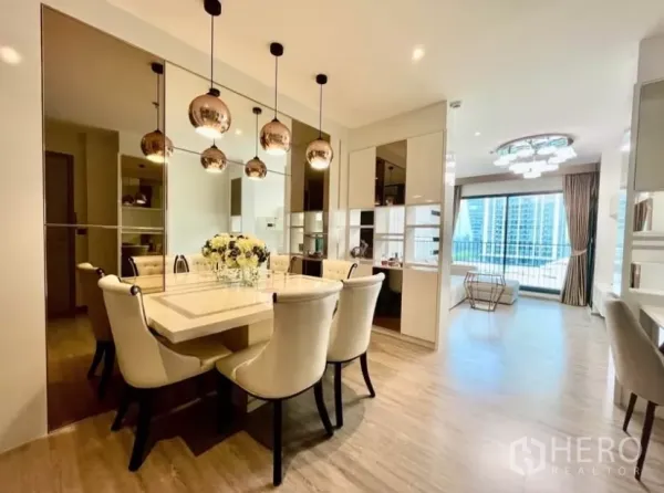 Condominium for rent in Watthana, Bangkok - Elegant dining area with mirrored wall, pendant lights and a six‑seat table opening to a bright living room and balcony city view.