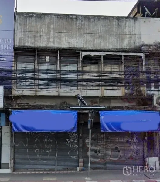 Shophouse for sale in Watthana, Bangkok - Street-facing shophouse facade with blue awnings, metal roller shutters, and upper barred windows in Watthana, Bangkok.