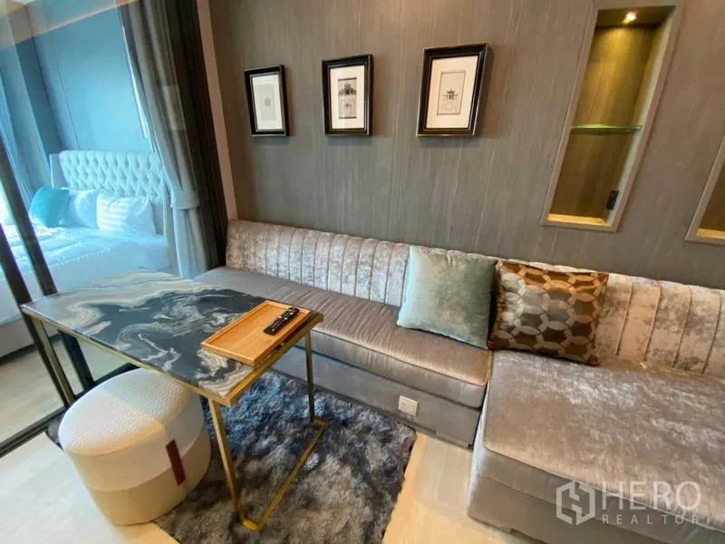 Condominium for rent in Pathum Wan, Bangkok - Stylish living room with long upholstered sofa, marble-look table and glass partition to bedroom.