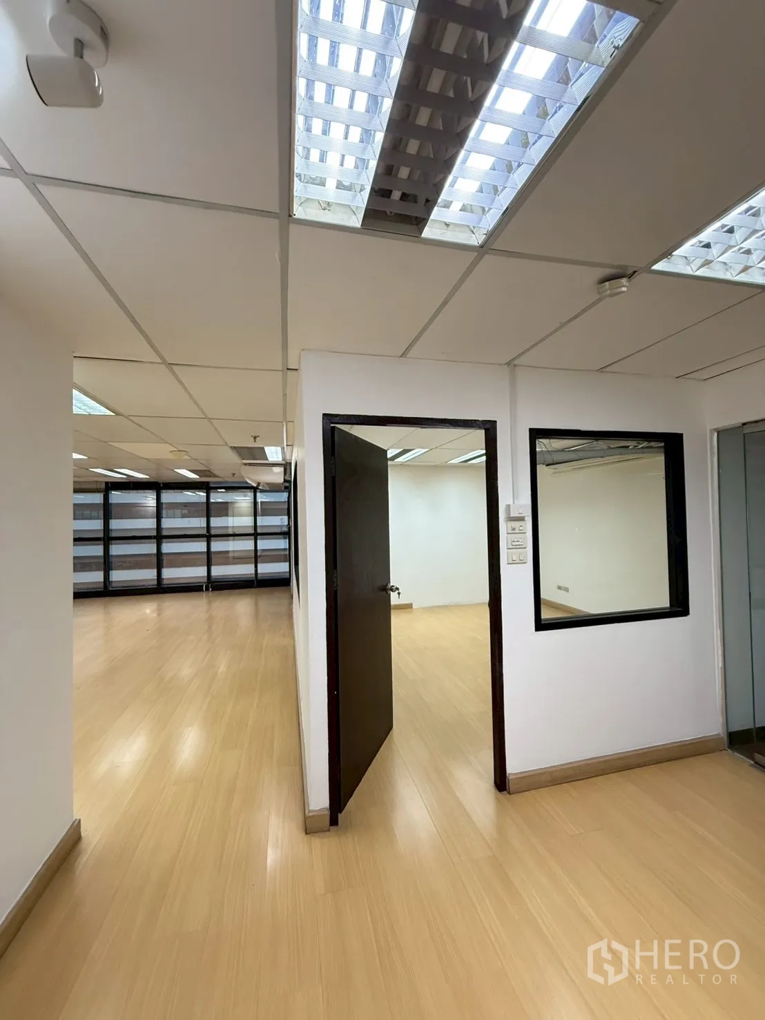 Office Space for rent in Watthana, Bangkok - Hallway to the open office with an internal glass window and dark wood door under fluorescent lighting.