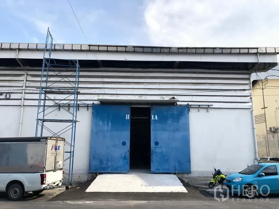 Warehouse for rent in Bang Phli, Samut Prakan - Warehouse exterior with white facade, wide blue sliding doors and a concrete loading ramp, with parking space in front.