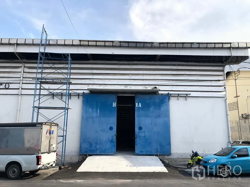 Warehouse for rent in Bang Phli, Samut Prakan - Warehouse exterior with white facade, wide blue sliding doors and a concrete loading ramp, with parking space in front.
