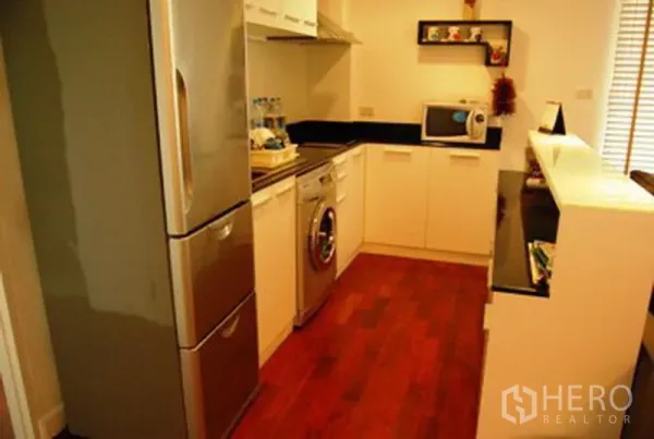 Condominium for rent in Pathum Wan, Bangkok - Compact U-shaped kitchen with stainless refrigerator, washing machine, white cabinetry and dark countertops.