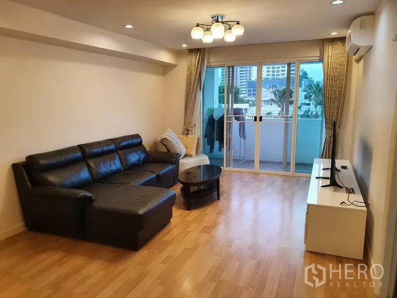 Condominium for rent in Watthana, Bangkok - Spacious living room with black leather sectional sofa, TV stand, and sliding doors to a balcony.