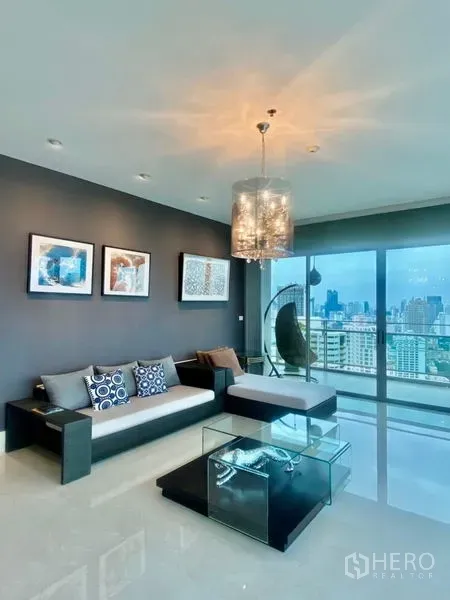 Condominium for rent in Watthana, Bangkok - Modern living room with chandelier, glass coffee table and floor-to-ceiling windows opening to a city-view balcony.