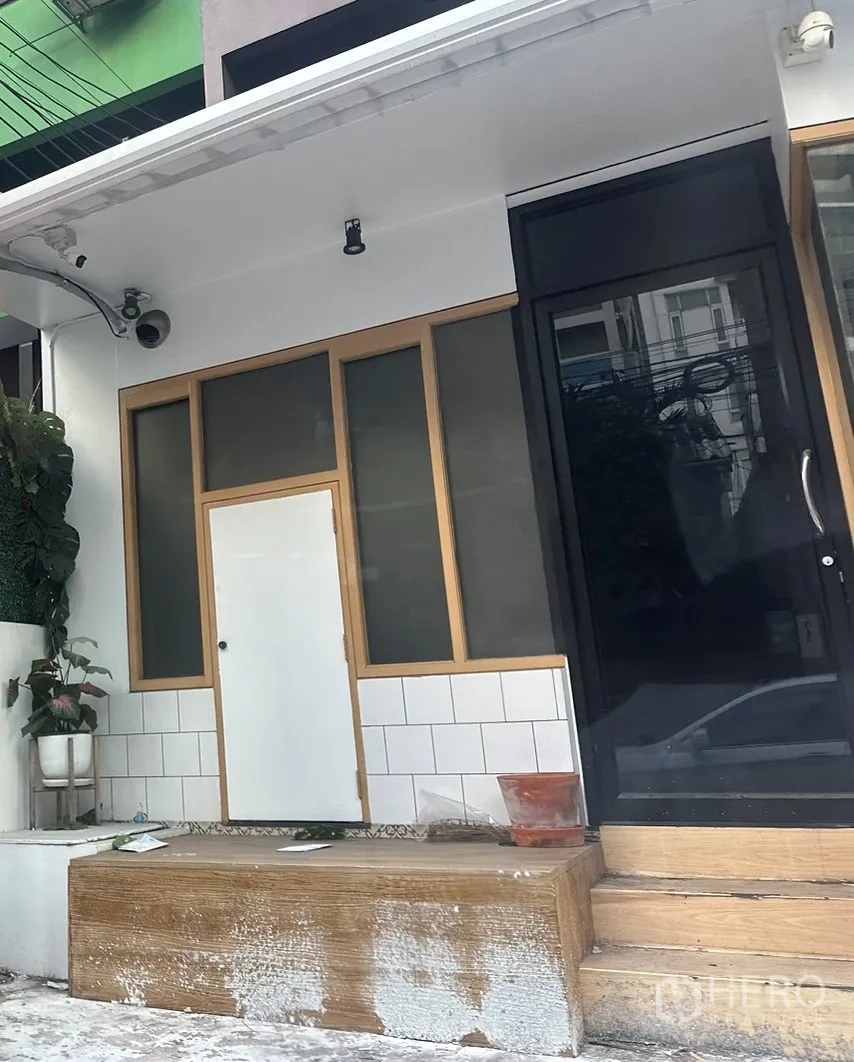 带办公室的家 出租在Watthana, Bangkok - 一层入口近景，黑色大门、磨砂窗、木质边框与监控摄像头。