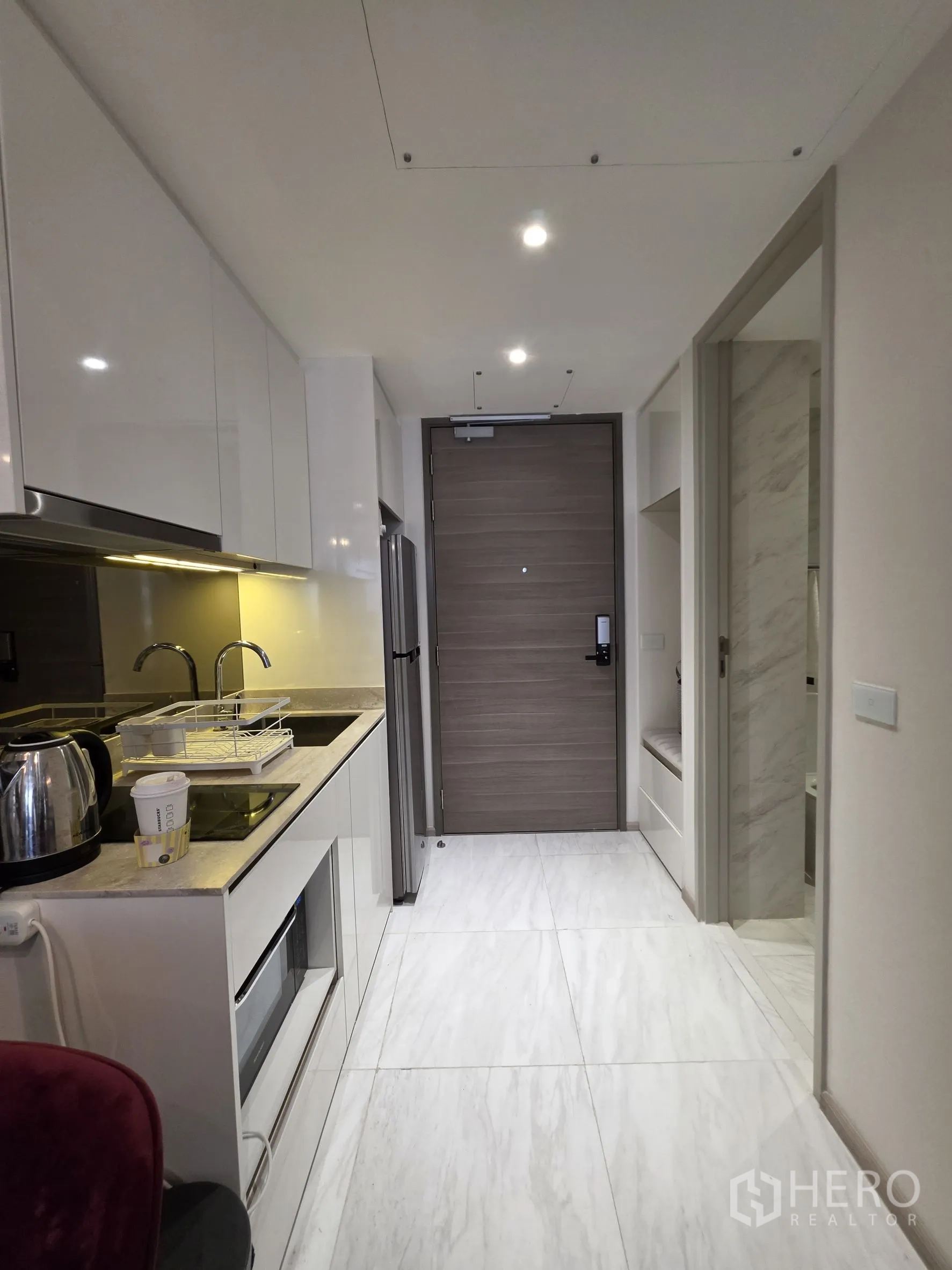Condominium for rent in Khlong Toei, Bangkok - Galley kitchen corridor with white cabinetry leading to the entrance; bathroom door on the right.