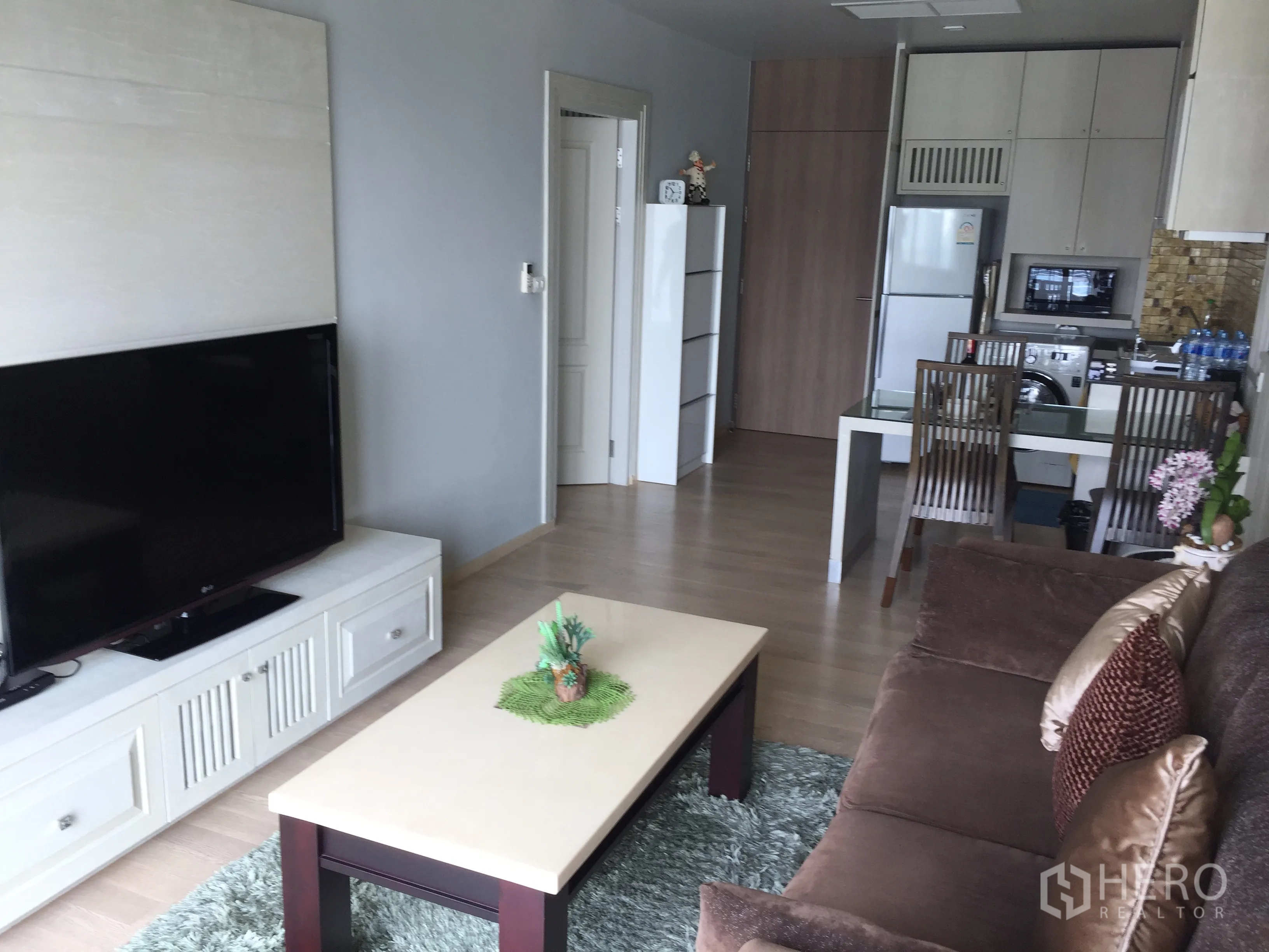 公寓 出租在Bangkok - 明亮客厅，配大屏电视、棕色沙发、茶几与开放式厨房餐区。