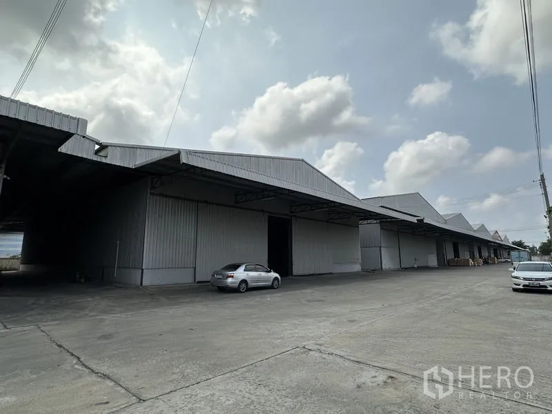 Warehouse for rent in Mueang Samut Prakan, Samut Prakan - Row of connected warehouse units with wide concrete apron and parked cars.