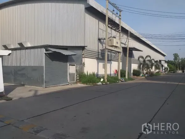 Factory for rent in Phra Nakhon Si Ayutthaya - Side view of a corrugated metal warehouse with a roller shutter door and utility lines along the street.