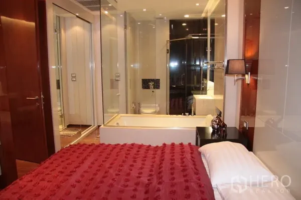 Condominium for rent in Khlong Toei, Bangkok - Bedroom with red duvet opening to a glass-walled bathroom featuring a bathtub and shower.