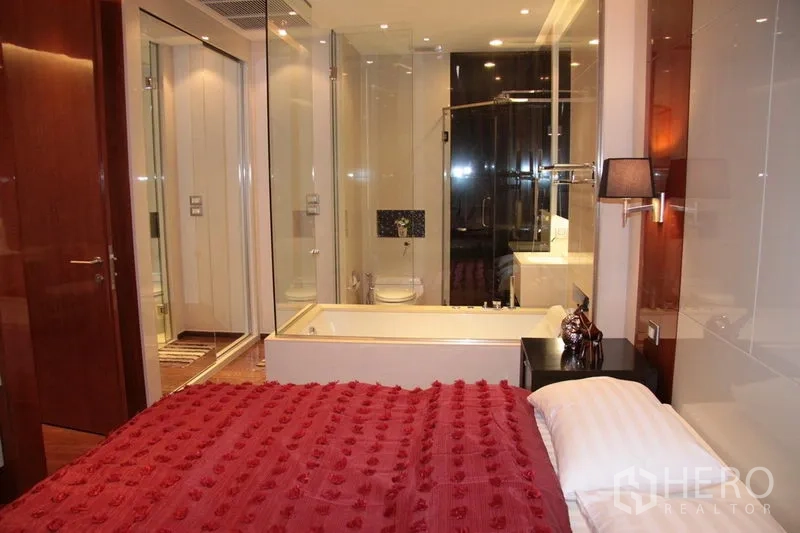 Condominium for rent in Khlong Toei, Bangkok - Bedroom with red duvet opening to a glass-walled bathroom featuring a bathtub and shower.