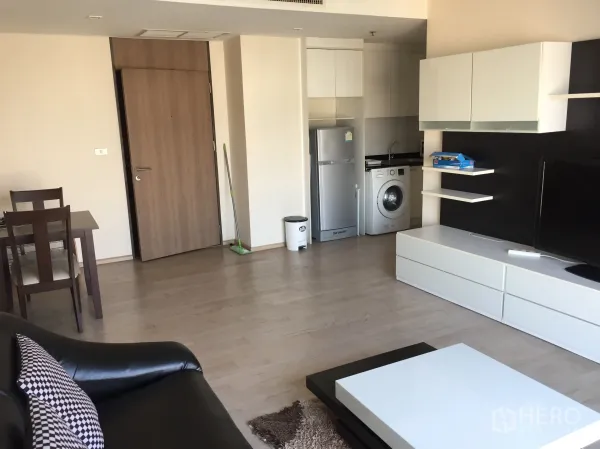 Condominium for rent or sale in Khlong Toei, Bangkok - Open-plan living room with dining set, TV console, and compact kitchenette with fridge and washing machine.