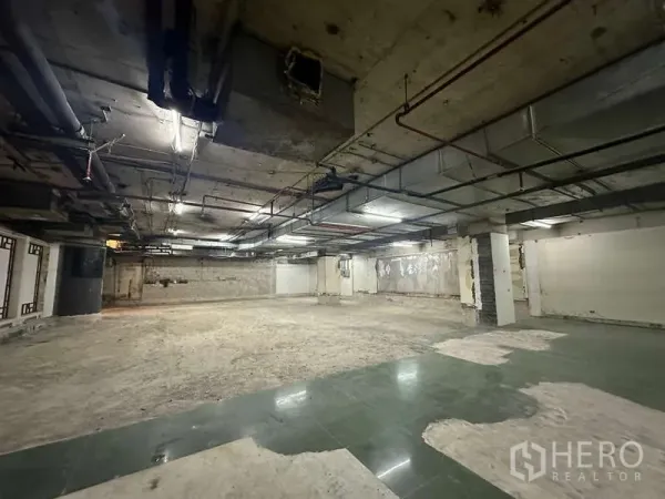 Retail Space for rent - Large unfinished retail floor with exposed ductwork, columns and partial green tile under fluorescent lighting.