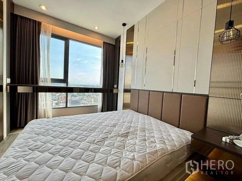 Condominium for rent or sale in Bang Na, Bangkok - Close view of the master bed with padded headboard next to a wide window and skyline backdrop.