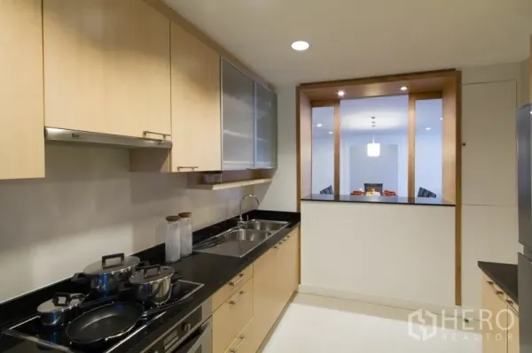 Apartment for rent in Watthana, Bangkok - Modern galley kitchen with light wood cabinetry, black granite countertops, double sink and a pass-through to the dining area.