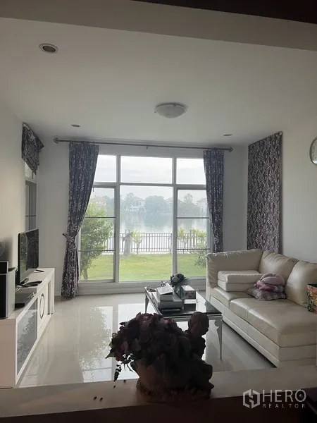 Detached House for rent or sale in Lat Krabang, Bangkok - Bright living room with white sofa and floor-to-ceiling windows facing a peaceful lakeside view.