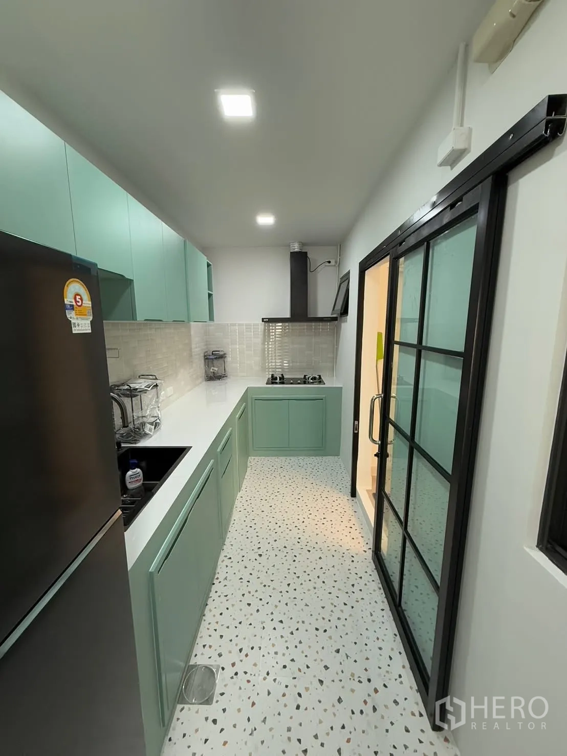 联排别墅 for rent in Bang Phli, Samut Prakan - 长形厨房配薄荷绿色橱柜、白色台面、燃气灶与水磨石地面，黑框推拉玻璃门分隔。