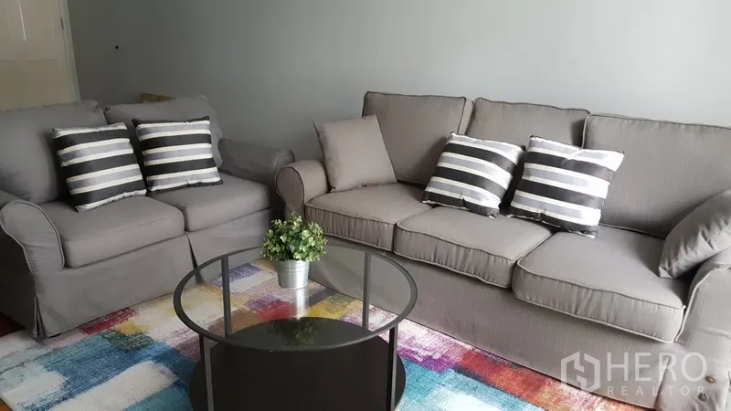 Condominium for rent in Watthana, Bangkok - Cozy living room with two gray sofas, striped cushions, a round glass coffee table, and a colorful rug.