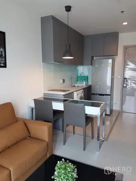 公寓 出租在Khlong Toei, Bangkok - 开放式小厨房，灰色橱柜、吊灯、餐桌、冰箱和洗衣机。