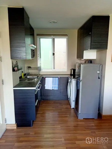 Condominium for rent in Khlong Toei, Bangkok - Galley kitchen with dark cabinets, window, sink, fridge and washing machine on wood floors.
