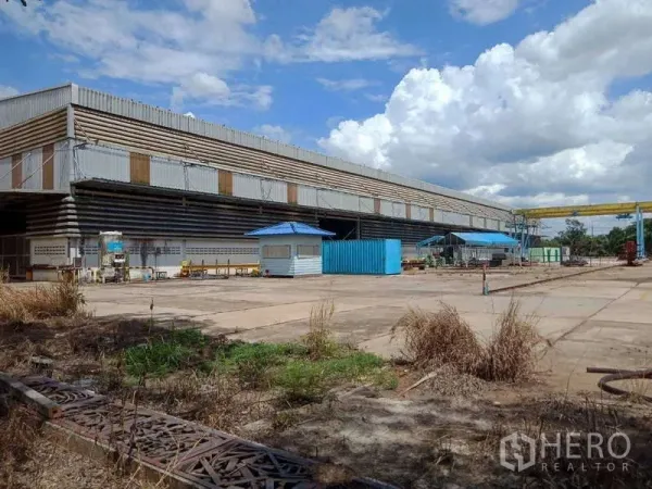 Factory for sale in Chachoengsao - Large steel-clad factory with a wide concrete yard, blue security booth, and gantry crane under cloudy skies.