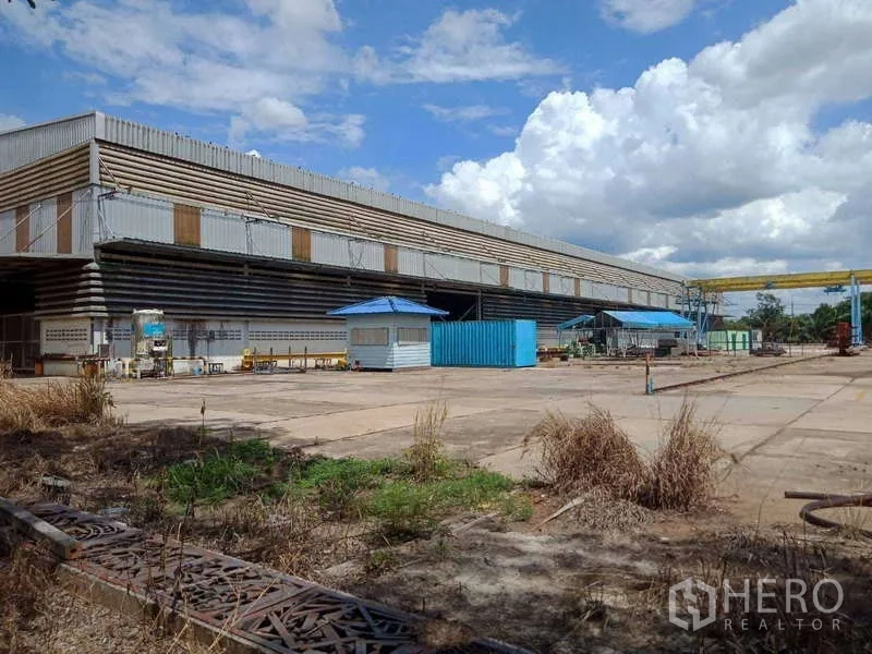 Factory for sale in Chachoengsao - Large steel-clad factory with a wide concrete yard, blue security booth, and gantry crane under cloudy skies.