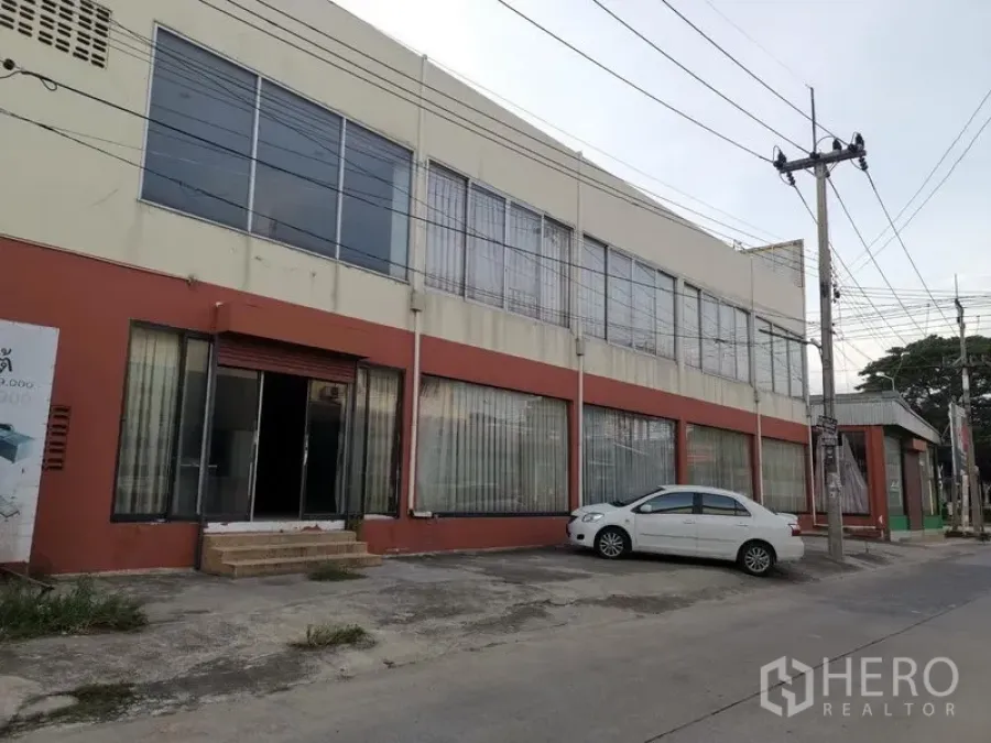 Retail Space for rent in Lam Luk Ka, Pathum Thani - Side facade of the commercial building showing tall glass windows, a roller door and small front parking area.