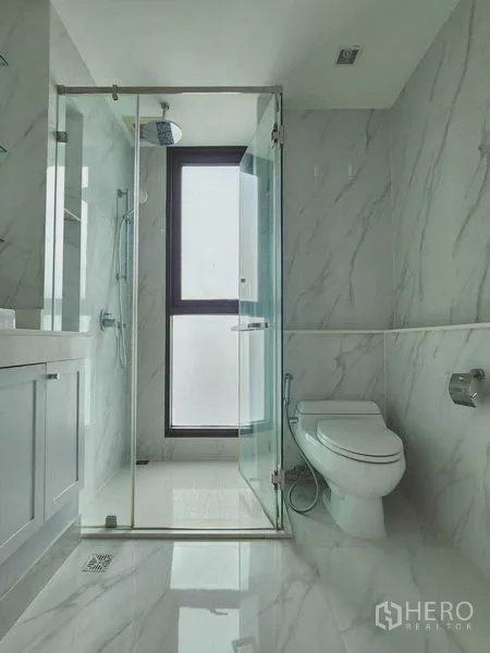 Condominium for rent in Bang Rak, Bangkok - Bright bathroom finished in white marble tiles with glass shower enclosure and window.