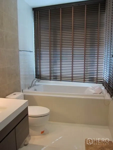 Condominium for rent in Khlong Toei, Bangkok - Modern bathroom with built-in bathtub, toilet and window blinds.