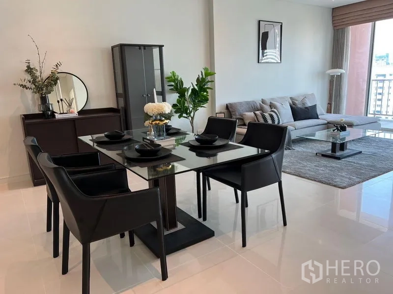 Condominium for rent in Watthana, Bangkok - Dining zone with a glass table and four black chairs beside a sofa and display cabinet.
