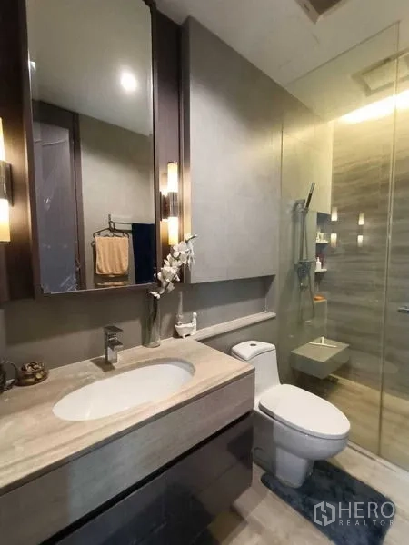 Condominium for rent or sale in Bang Rak, Bangkok - Contemporary bathroom with marble-top vanity, toilet and glass-enclosed shower with warm lighting.