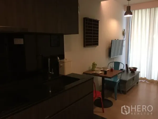 Condominium for rent in Watthana, Bangkok - Compact kitchen with dark cabinets, sink, pendant light and a small dining table facing a wall-mounted TV.