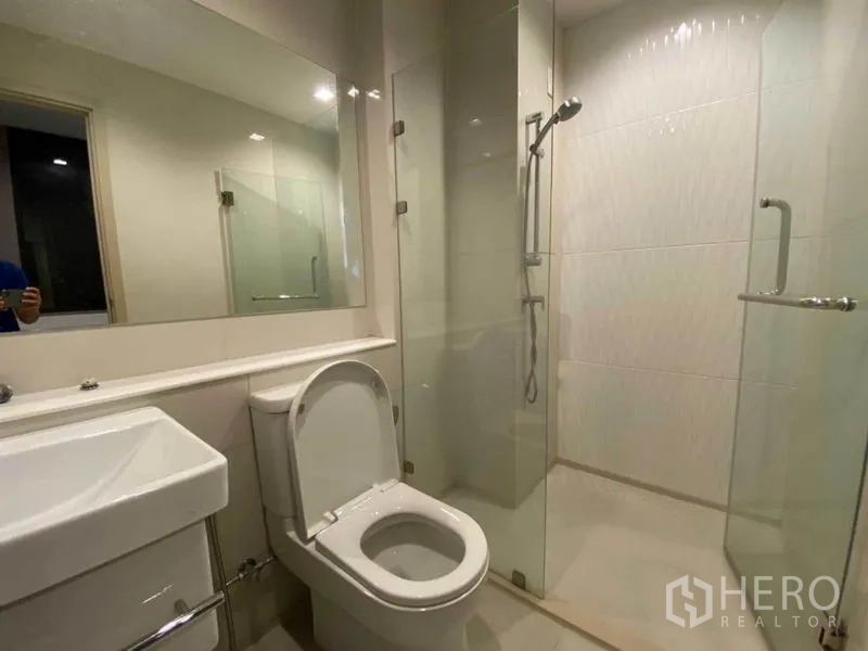 Condominium for rent in Pathum Wan, Bangkok - Modern bathroom with a large mirror, toilet and a glass-enclosed shower.