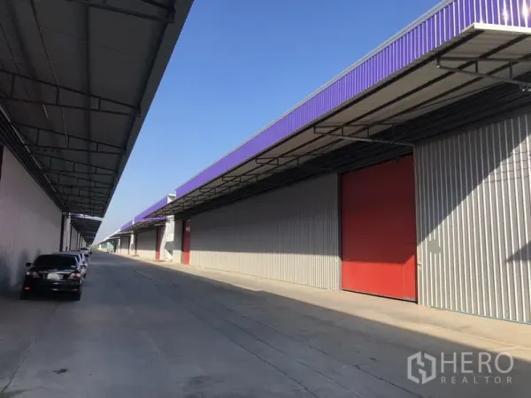 Warehouse for rent in Pathum Thani - Row of modern warehouses with purple eaves and large red sliding doors along a wide driveway.