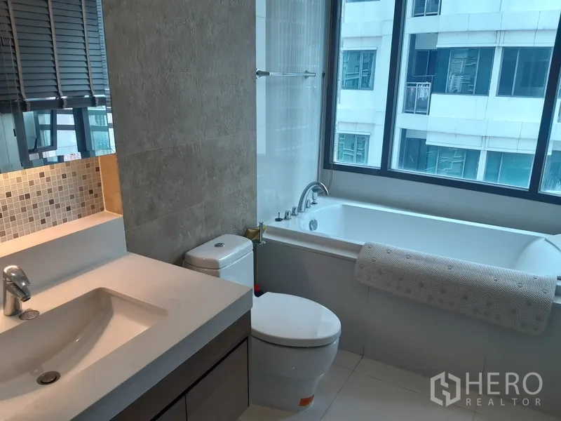 Condominium for rent in Khlong Toei, Bangkok - Contemporary bathroom with bathtub by the window, toilet, and vanity with mosaic backsplash.