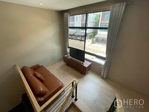 Townhouse for rent in Suan Luang, Bangkok - Upper lounge with brown leather sofa, TV stand and a wide window overlooking a quiet residential lane.