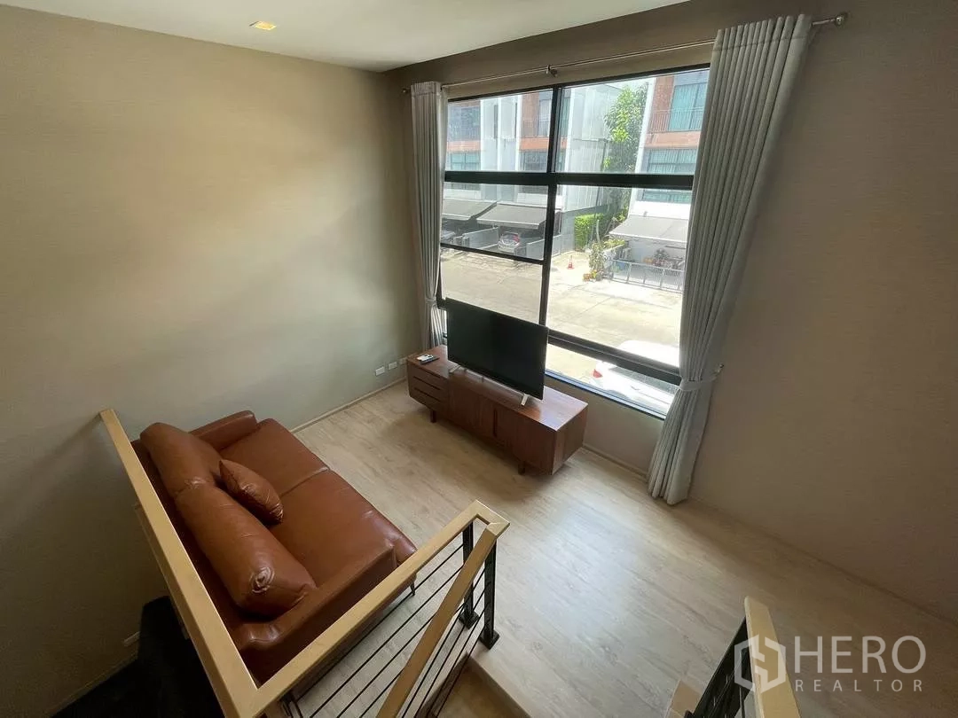 Townhouse for rent in Suan Luang, Bangkok - Upper lounge with brown leather sofa, TV stand and a wide window overlooking a quiet residential lane.