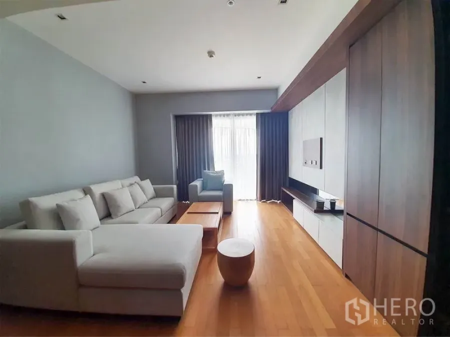 公寓 出租在Yan Nawa, Bangkok - 明亮客厅，L形沙发、木地板与电视墙，通向带窗帘的阳台。