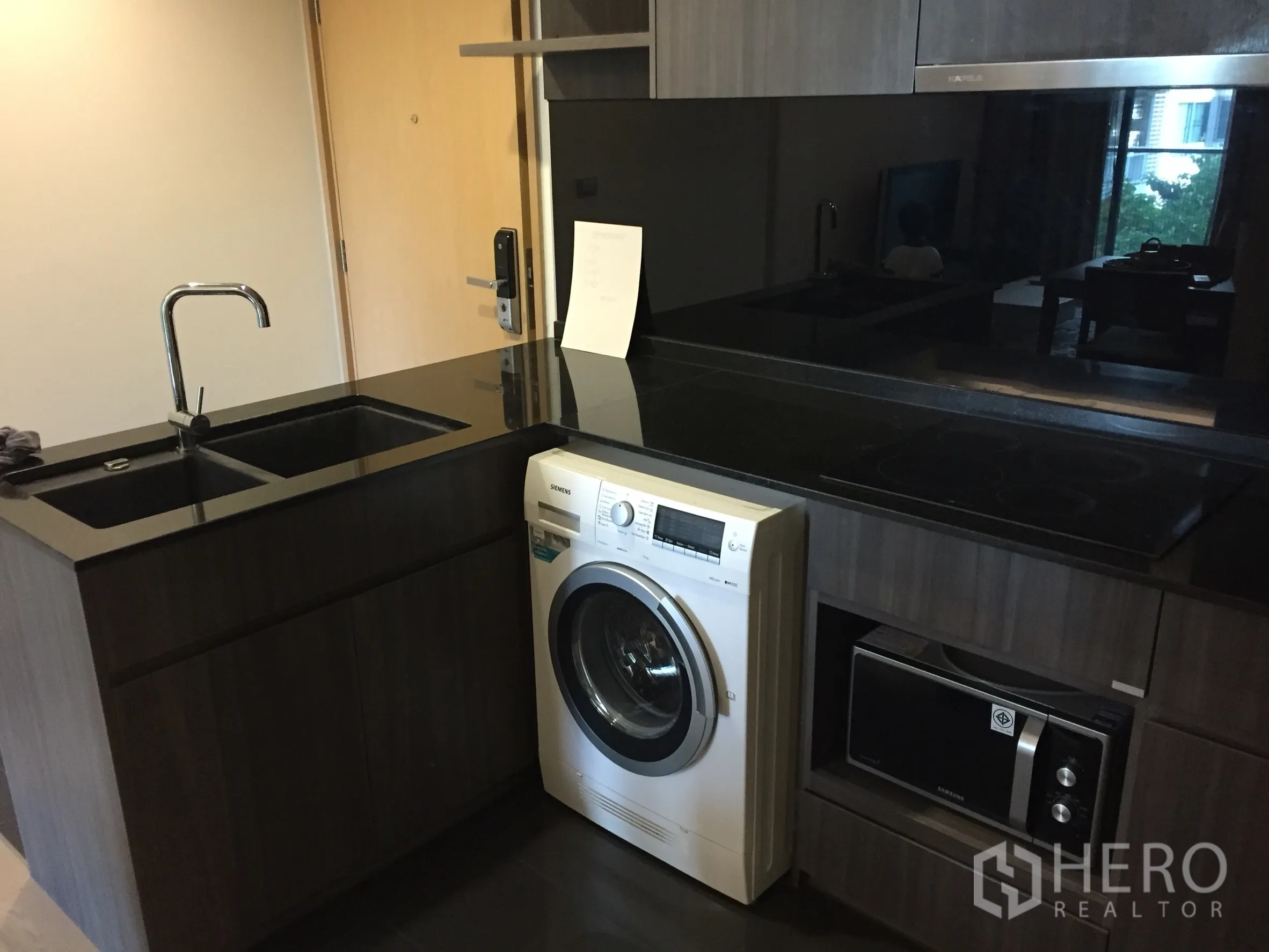 Condominium for rent or sale in Watthana, Bangkok - Modern compact kitchen with black granite counters, double sink, built-in cooktop, washing machine, and microwave in dark wood cabinetry.